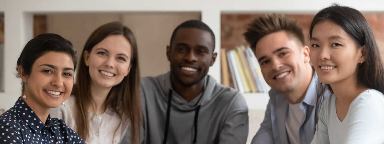 Group picture of happy multiethnic young people sit at shared desk look at camera studying together, multicultural excited students or groupmates smiling posing for photo working in library